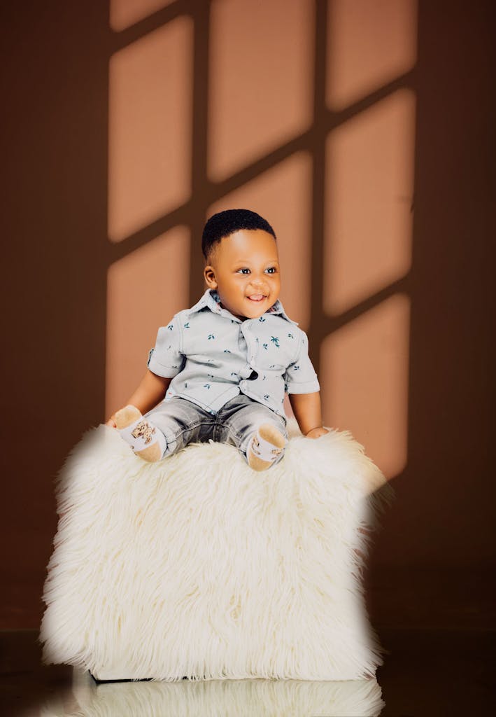 Charming baby boy in casual attire sitting on a fluffy stool against a warm backdrop.