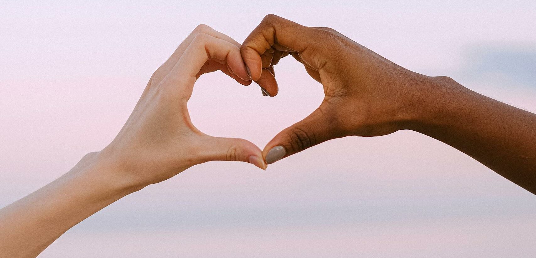 A powerful image of unity and diversity with two hands forming a heart against a serene pastel sky.